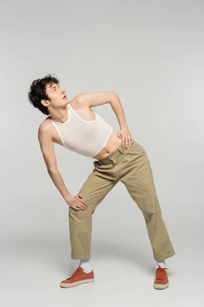 Full length of trendy pansexual person in crop top and beige pants looking away on grey background — Stock Photo