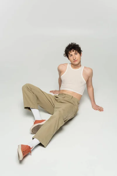 Longitud completa del modelo pangender en top de recorte y pantalones beige sentados sobre fondo gris - foto de stock