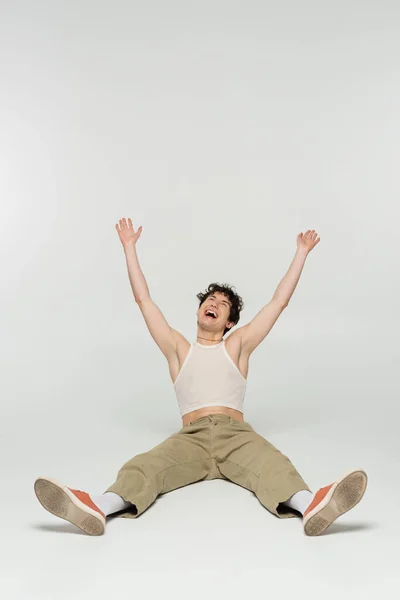 Excited nonbinary person in beige pants laughing while sitting with raised hands on grey background — Stock Photo