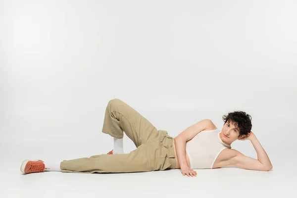 Full length of young pansexual person in stylish clothes lying and looking at camera on grey background — Stock Photo