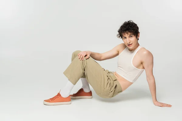 Full length of brunette pansexual model in beige pants looking at camera while posing on grey background — Stock Photo