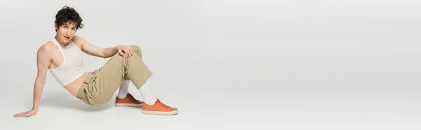 Полная длина модной модели pangender в бежевых брюках и резиновых туфлях, смотрящих в камеру, позируя на сером фоне, баннер — стоковое фото