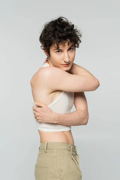Brunette nonbinary model in crop top hugging own body and looking at camera isolated on grey — Stock Photo