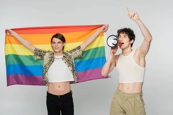 Joven persona no binaria gritando en megáfono cerca de socio positivo con bandera lgbt aislado en gris - foto de stock