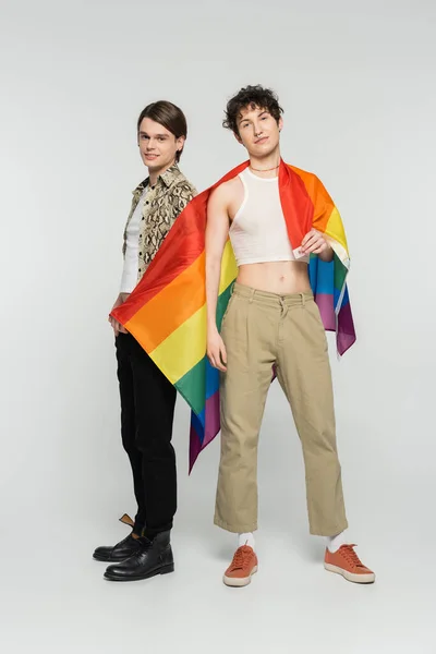 Full length of trendy pansexual partners with lgbt flag looking at camera while standing on grey background — Stock Photo