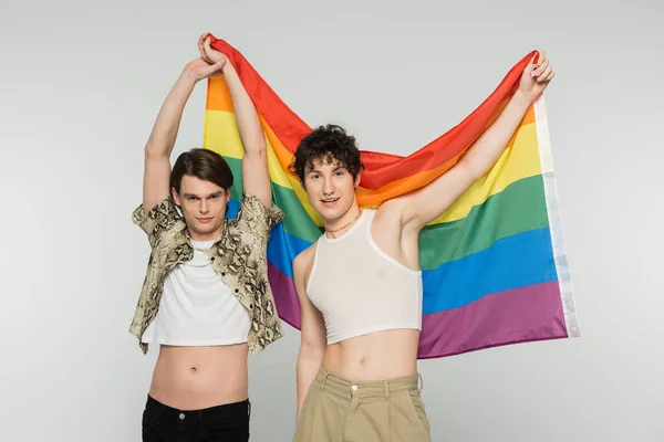 Positive nonbinary couple in stylish clothes holding rainbow flag and looking at camera isolated on grey — Stock Photo