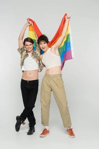 Full length of joyful bigender partners holding lgbt flag in raised hands on grey background — Stock Photo