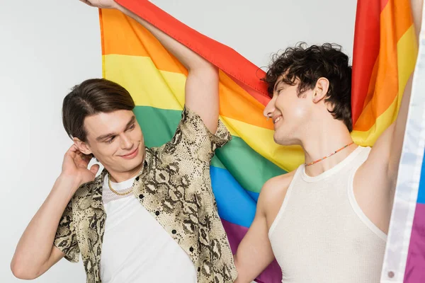 Cheerful nonbinary couple holding lgbt flag and smiling at each other isolated on grey — Stock Photo