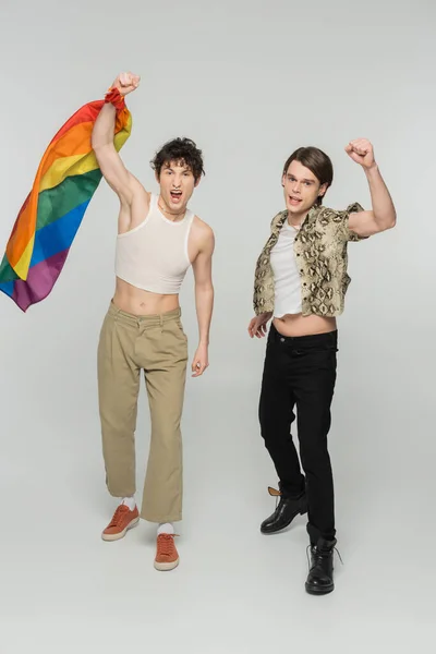 Excited pangender partners with lgbt flag gesturing with clenched fists and screaming on grey background — Stock Photo