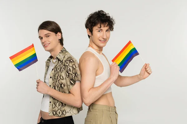 Moda e despreocupado casal pansexual de pé com pequenas bandeiras lgbt e sorrindo para a câmera isolada em cinza — Fotografia de Stock