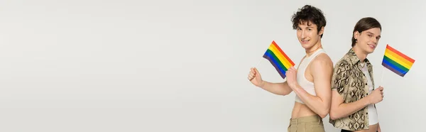 Happy nonbinary couple holding small lgbt flags and smiling at camera isolated on grey, banner — Stock Photo