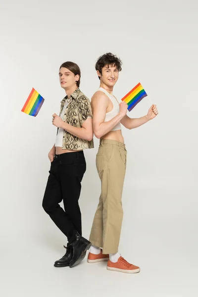 Full length of stylish pansexual models posing with small lgbt flags on grey background — Stock Photo