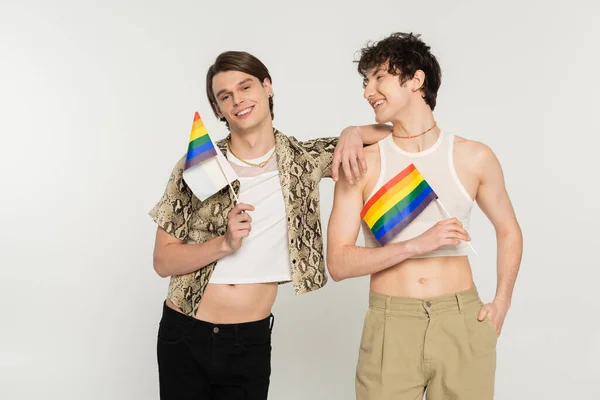 Cheerful and stylish bigender models posing with small lgbt flags isolated on grey — Stock Photo