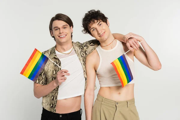 Pareja grande joven y despreocupada con banderas de arco iris abrazando y sonriendo a la cámara aislada en gris - foto de stock