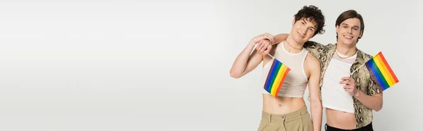 Young and carefree pangender partners holding small lgbt flags and smiling at camera isolated on grey, banner — Stock Photo