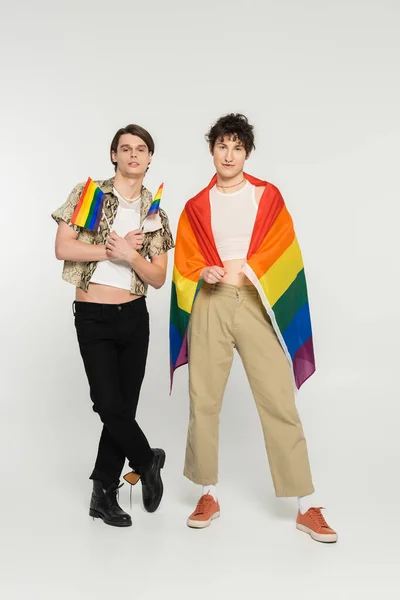 Comprimento total de jovens modelos não binários em roupas elegantes posando com bandeiras de arco-íris em fundo cinza — Fotografia de Stock
