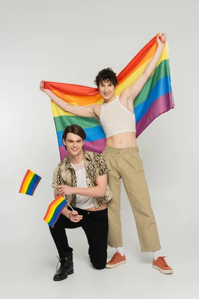 Piena lunghezza della coppia pansessuale positiva che tiene bandiere arcobaleno mentre guarda la fotocamera su sfondo grigio — Foto stock