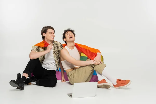Excited pangender couple with rainbow flag sitting near laptop on grey background — Stock Photo