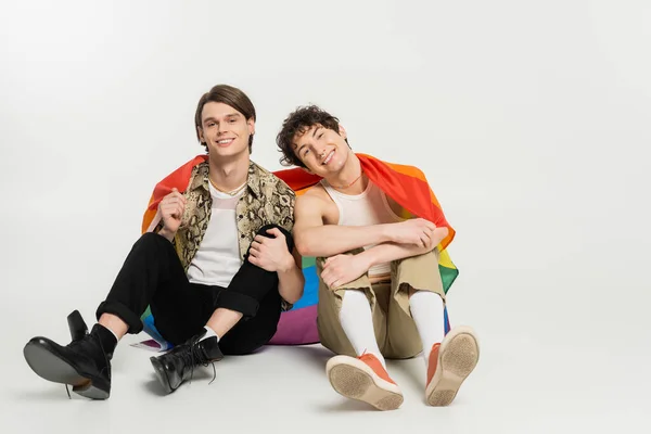 Full length of trendy and joyful nonbinary partners sitting with rainbow flag on grey background — Stock Photo