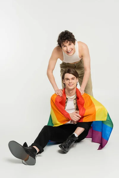 Persona no binaria llena de alegría sonriendo cerca de su pareja sentada con la bandera del arco iris sobre fondo gris - foto de stock