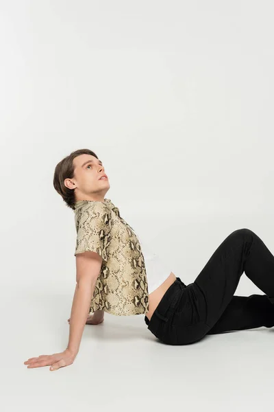 Vista lateral de la joven persona pansexual en ropa de moda mirando hacia arriba mientras está sentado sobre fondo gris - foto de stock