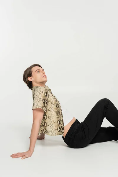 Side view of young pansexual person in trendy clothes looking up while sitting on grey background — Stock Photo