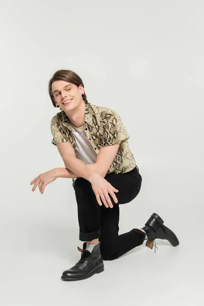 Full length of cheerful nonbinary model in black pants and animal print blouse looking away on grey background — Stock Photo