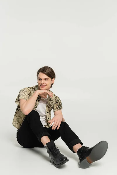 Full length of carefree bigender person in black trousers and snakeskin print blouse sitting on grey background — Stock Photo