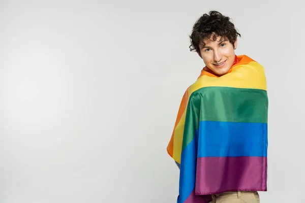 Modelo pansexual feliz cubierto con la bandera del arco iris sonriendo a la cámara aislada en gris - foto de stock