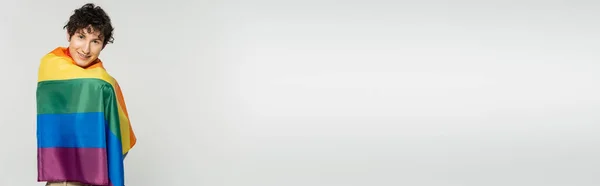 Felice persona non binaria con i capelli castani ondulati in piedi sotto bandiera lgbt isolato su grigio, banner — Stock Photo