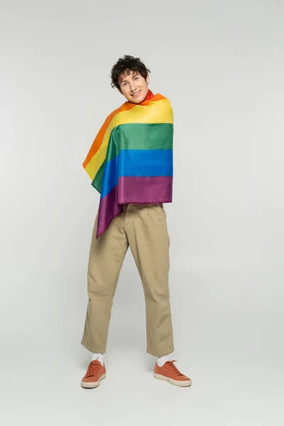 Full length of smiling bigender person in beige pants posing with rainbow flag on grey background — Stock Photo