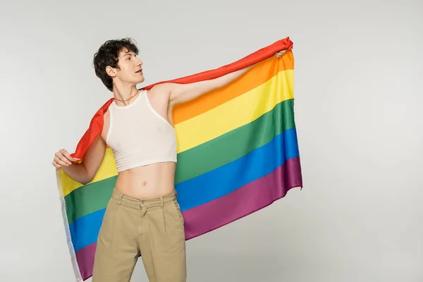 Stylish nonbinary person in crop top and pants posing with rainbow flag isolated on grey — Stock Photo