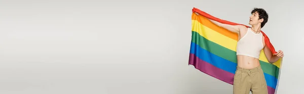 Brunette nonbinary model in crop top posing with lgbt flag isolated on grey, banner — Stock Photo