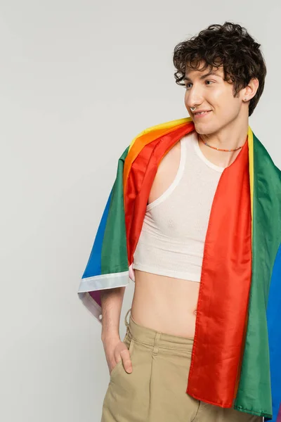 Carefree nonbinary person with lgbt flag on shoulders looking away and smiling isolated on grey — Stock Photo