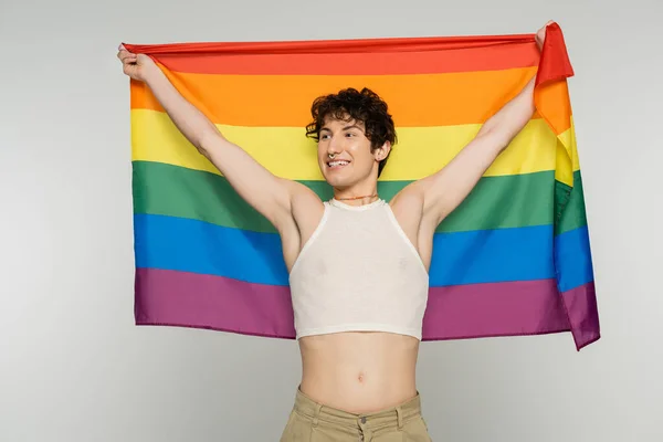 Joyful bigender model in crop top holding lgbt flag in raised hands and looking away isolated on grey — Stock Photo