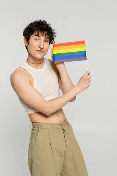 Cheerful brunette pansexual model in crop top showing small lgbt flag isolated on grey — Stock Photo