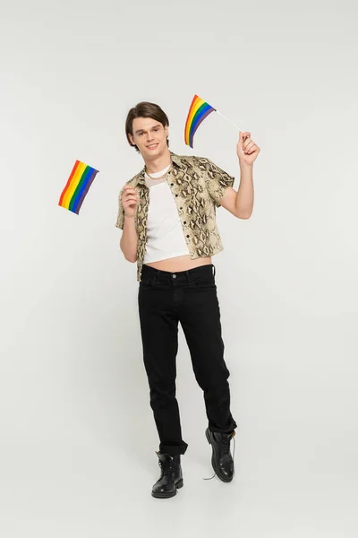 Full length of positive pansexual model in black pants and snakeskin print blouse holding small lgbt flags on grey background — Stock Photo
