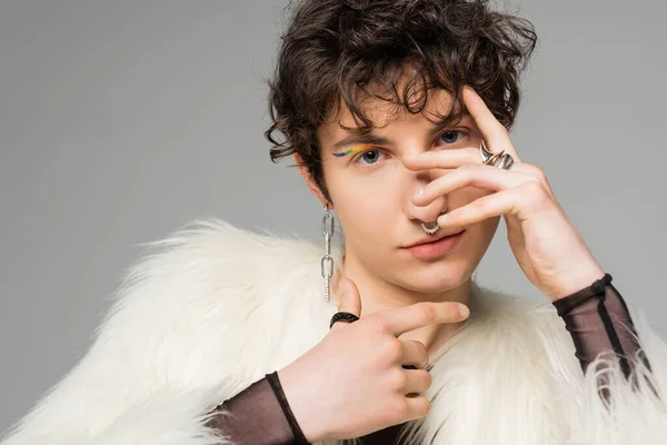 Brunette pansexual person in white faux fur jacket and silver accessories obscuring face with hand isolated on grey — Stock Photo