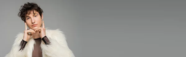 Persona alla moda bigender in giacca di eco pelliccia bianca tenendosi per mano vicino al viso e guardando la fotocamera isolata su grigio, banner — Foto stock