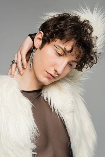 Portrait of fashionable bigender person with rainbow eye liner posing in white faux fur jacket isolated on grey — Stock Photo