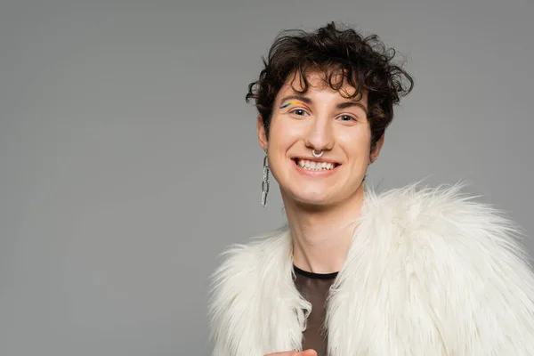 Overjoyed pansexual model in silver accessories and white faux fur jacket smiling at camera isolated on grey — Stock Photo