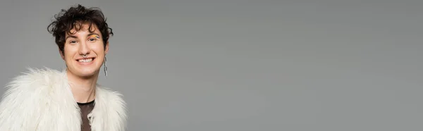 Fröhliche bigeschlechtliche Person in weißer Kunstpelzjacke und silbernem Ohrring isoliert auf grau, Banner — Stockfoto