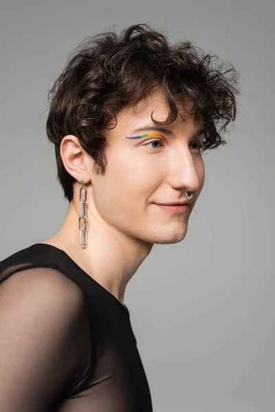 Porträt eines lächelnden Bigender-Modells mit Regenbogen-Eyeliner und silbernen Accessoires isoliert auf grau — Stockfoto