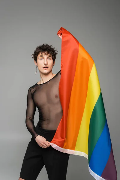 Young bigender model in black transparent top holding lgbt flag isolated on grey — Stock Photo