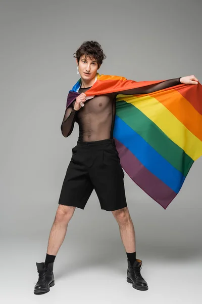 Full length of brunette nonbinary model in black shorts and leather boots posing with lgbt flag on grey background — Stock Photo