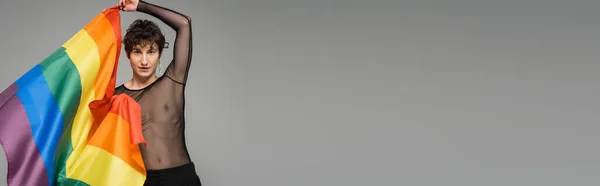 Bruna bigender persona in nero trasparente top guardando la fotocamera mentre tenendo lgbt bandiera isolata su grigio, banner — Foto stock