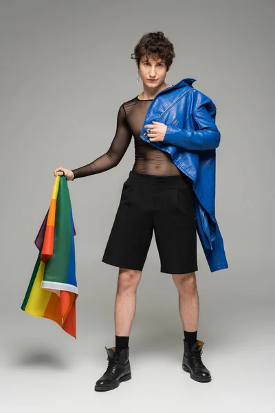 Full length of trendy pangender person with blue leather jacket holding lgbt flag on grey background — Stock Photo