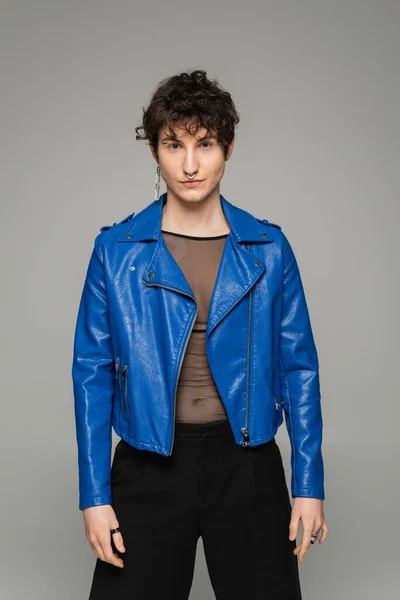 Модная пансексуальная модель в синей кожаной куртке, смотрящая на камеру, изолированную на сером — стоковое фото