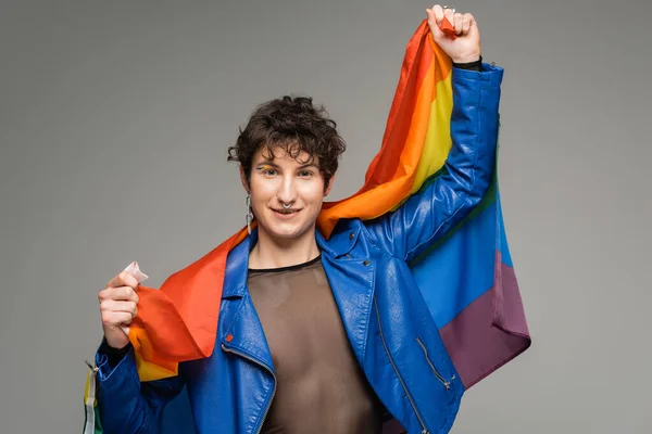 Persona pangender spensierata in giacca di pelle blu con bandiera arcobaleno e guardando la fotocamera isolata sul grigio — Foto stock