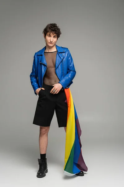 Longitud completa de la persona pansexual con bandera lgbt posando en chaqueta de cuero azul y pantalones cortos negros sobre fondo gris - foto de stock